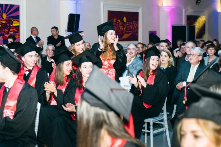 Un groupe de diplômés en toge noire et casquette avec étole rouge sont assis et debout en rangs lors de la cérémonie de remise des diplômes de l'EBBS. Le public derrière eux, composé d'hommes et de femmes de différents âges, applaudit. La salle est décorée d'œuvres d'art murales colorées et illuminée par un éclairage doux.