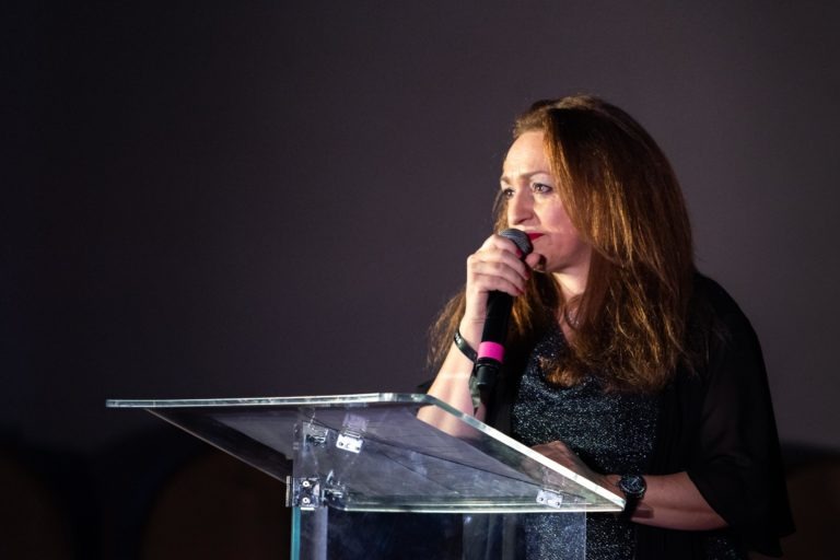 Lors de la cérémonie de remise des diplômes EBBS, une femme aux longs cheveux bruns parle dans un micro, debout derrière un podium transparent. Elle porte une tenue noire et est éclairée par un projecteur, sur fond sombre et flou. Sa main gauche repose sur le podium tandis qu'elle tient le micro dans sa main droite.