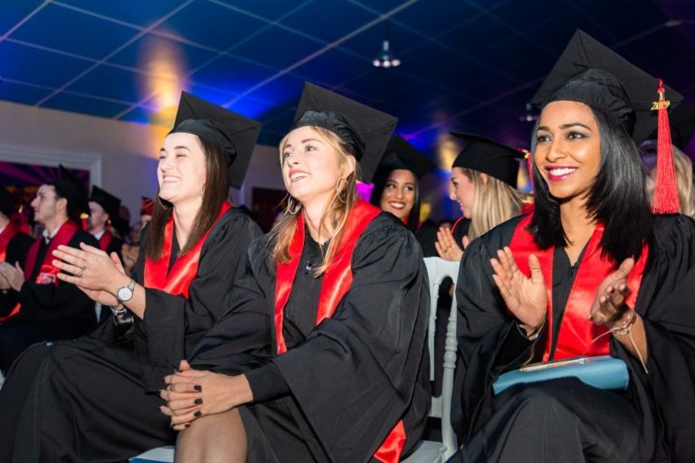 Un groupe de diplômés portant des toges et des casquettes noires avec des étoles rouges sont assis en rang, applaudissant et souriant. L'arrière-plan montre une salle avec un plafond bleu et des lumières colorées. L'atmosphère de la cérémonie de remise des diplômes EBBS est animée et festive.