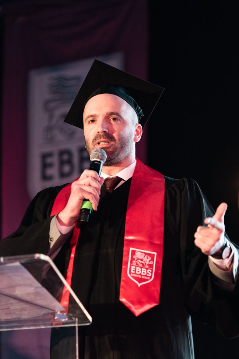 Un homme en tenue de remise de diplômes, avec une robe noire, une casquette et une étole rouge avec le logo de l'EBBS, parle dans un microphone sur un podium lors de la cérémonie de remise des diplômes EBBS. Il fait un geste d'une main tout en tenant le microphone de l'autre. Une bannière affichant le logo EBBS est visible en arrière-plan.