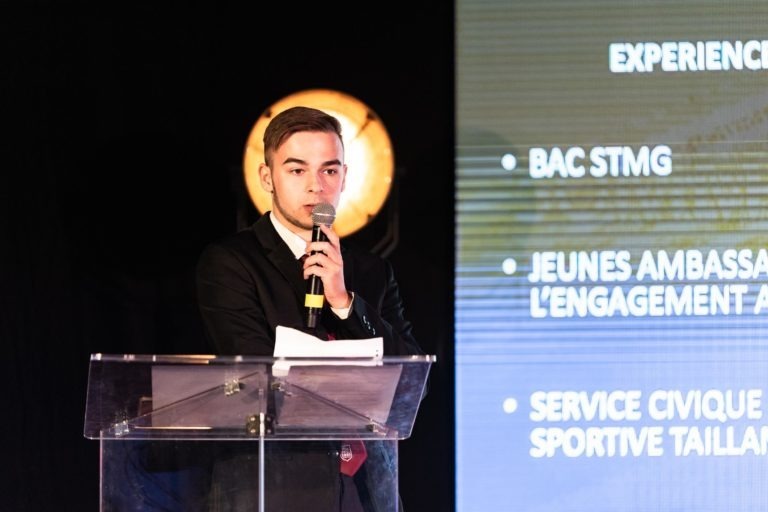 Un jeune homme en costume sombre et cravate rouge se tient debout sur un podium dégagé, tenant un micro et parlant. Derrière lui, un écran de présentation affiche un texte en français, énumérant des éléments tels que « BAC STMG », « Jeunes Ambassadeur » et « Service Civique Sportif Taill ». L'arrière-plan fait allusion à la prestigieuse Cérémonie de remise des diplômes EBBS.