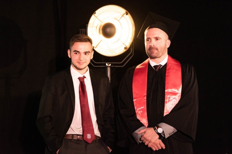 Deux hommes se tiennent côte à côte sur un fond sombre avec une lumière circulaire brillante derrière eux. L'homme de gauche porte un costume noir, une chemise blanche et une cravate rouge. L'homme de droite porte une toge et une toge de remise de diplôme avec une ceinture rouge, marquant la cérémonie de remise des diplômes de l'EBBS. Tous deux ont une expression sérieuse.