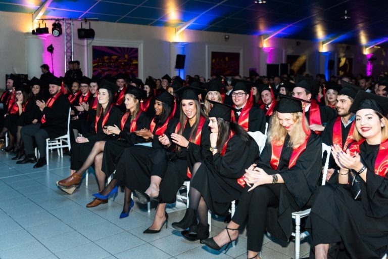 Un grand groupe d'étudiants vêtus de toges noires et de casquettes avec des étoles rouges sont assis sur des rangées de chaises, applaudissant et souriant lors d'une cérémonie de remise de diplômes EBBS en salle. La salle est éclairée de lumières colorées et l'atmosphère est festive. Certains étudiants ont les jambes croisées tandis que d'autres applaudissent avec enthousiasme.