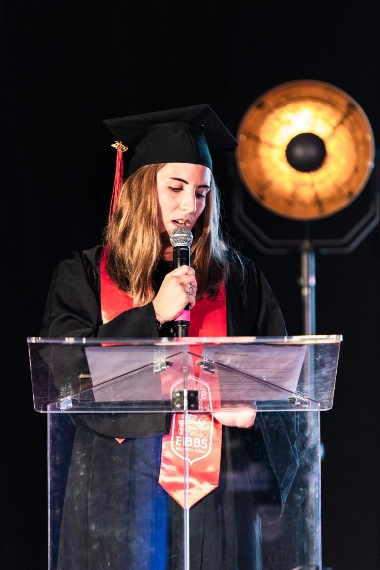 Une personne portant une toge et un chapeau noirs avec une étole rouge se tient debout sur un podium transparent, parlant dans un microphone pendant la cérémonie de remise des diplômes de l'EBBS. Derrière elle, une grande lumière ronde de studio est allumée, projetant une lueur chaleureuse. Le fond sombre met en valeur l'orateur et la lumière.