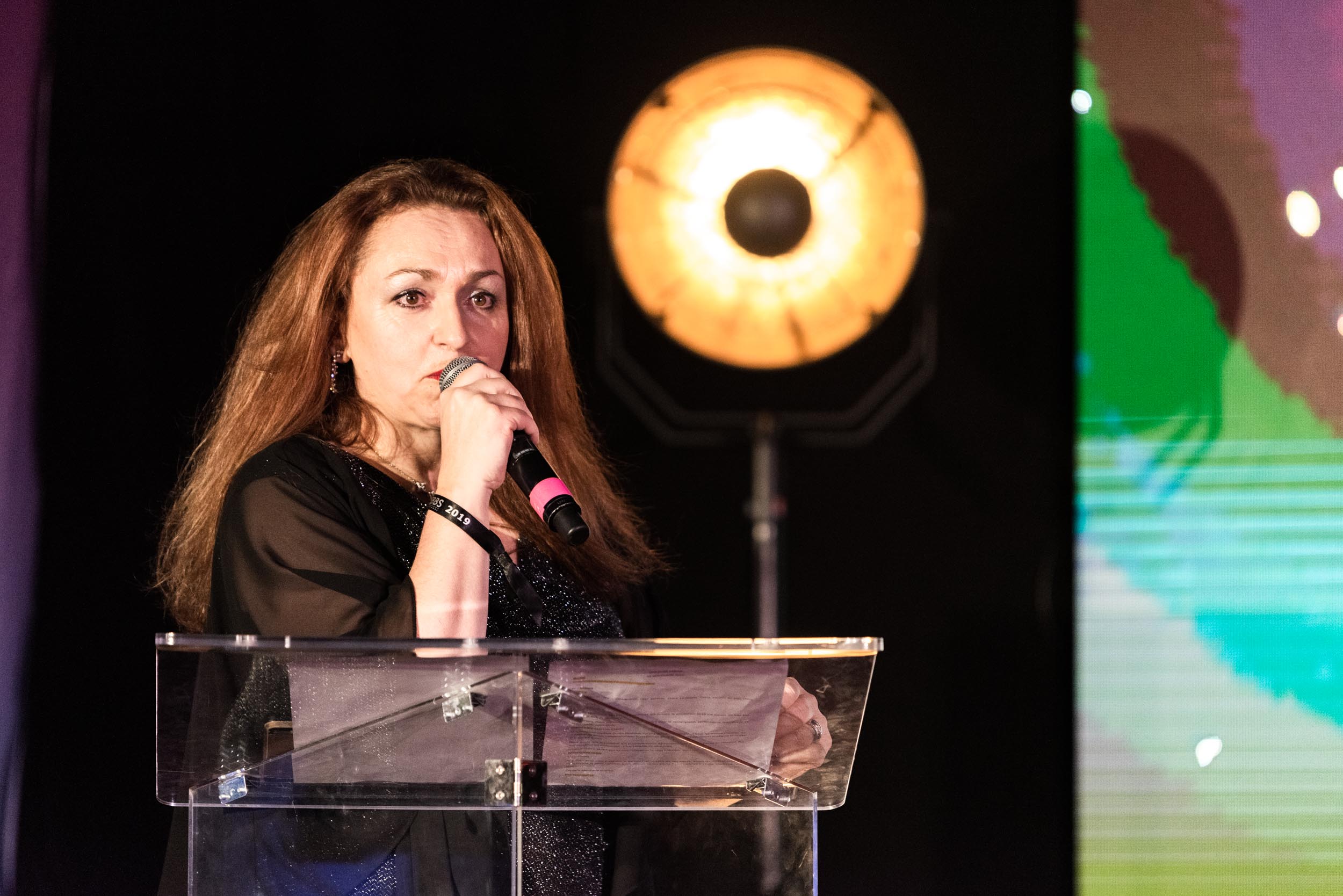 Lors de la cérémonie de remise des diplômes EBBS, une femme aux cheveux longs et bruns parle dans un micro, debout derrière un podium transparent. Elle porte une tenue sombre et se tient devant une lumière vive qui crée une lueur forte et chaleureuse. L'arrière-plan comprend un éclairage coloré et abstrait.