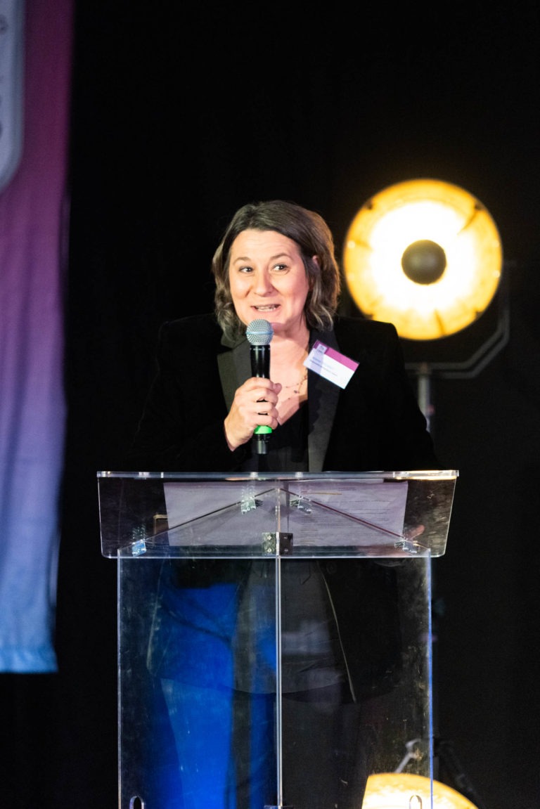 Une personne se tient derrière un podium transparent, tient un micro et parle lors de la cérémonie de remise des diplômes EBBS. Elle a les cheveux mi-longs et porte une veste sombre. Une lumière de scène ronde est allumée derrière elle, mettant en valeur la bannière partiellement visible avec des couleurs dégradées en arrière-plan.