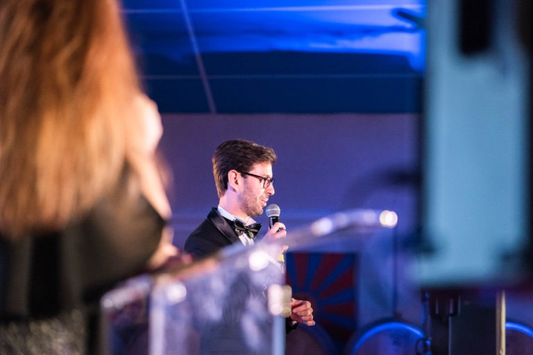 Un homme en costume et nœud papillon se tient debout sur un podium, tenant un microphone devant sa bouche pendant qu'il parle. Le décor semble être celui d'un événement en salle avec un éclairage ambiant bleu, probablement la cérémonie de remise des diplômes EBBS. L'arrière-plan montre une partie d'un plafond coloré et une silhouette floue au premier plan, vêtue de noir.