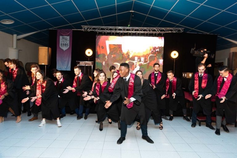 Lors de la cérémonie de remise des diplômes EBBS, un groupe d'étudiants en toge noire et étole rouge ont exécuté une chorégraphie sur scène. Disposés en rangs, ils ont dansé devant un écran vibrant affichant une toile de fond colorée, sous un plafond en carrelage bleu et un éclairage professionnel.