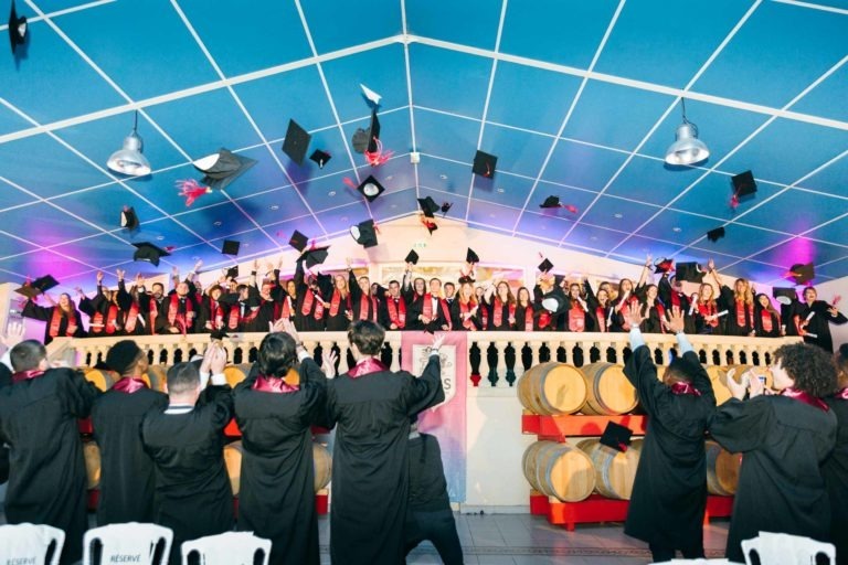 Un grand groupe de diplômés vêtus de toges noires et de casquettes célèbrent leur diplôme en lançant leurs casquettes en l'air. Ils se tiennent debout sur une scène sous un plafond grillagé bleu. Au premier plan, certaines personnes tournées vers la scène immortalisent l'instant avec leurs téléphones. La scène, qui fait partie de la cérémonie de remise des diplômes EBBS, est ornée de gros tonneaux.