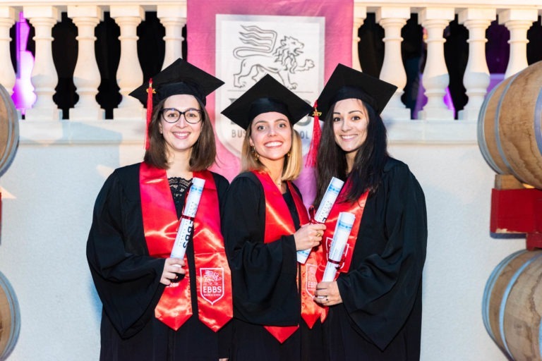 Trois femmes en toge noire et casquette de remise de diplômes se tiennent debout, souriantes, devant une balustrade blanche et une bannière sur laquelle est inscrit « EBBS » avec un emblème de lion. Elles tiennent chacune un diplôme enroulé et portent des écharpes rouges, symbolisant leur réussite lors de la cérémonie de remise des diplômes EBBS. L'arrière-plan est doucement éclairé, renforçant l'ambiance festive.