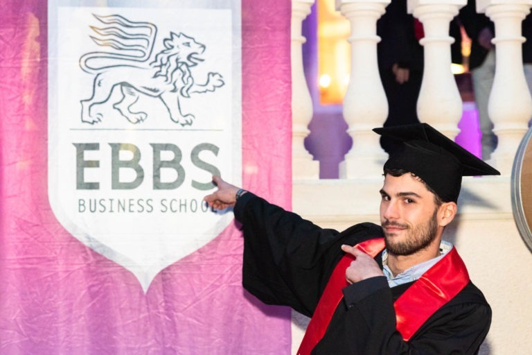 Un diplômé, vêtu d'une toge et d'une casquette noires bordées de rouge, pointe du doigt une grande bannière portant le logo de l'EBBS Business School. La bannière, qui présente un emblème de lion et le nom de l'école en lettres grasses, ajoute du sérieux à la cérémonie de remise des diplômes EBBS. L'événement officiel se déroule sur un arrière-plan flou avec des lumières vives et des détails architecturaux.