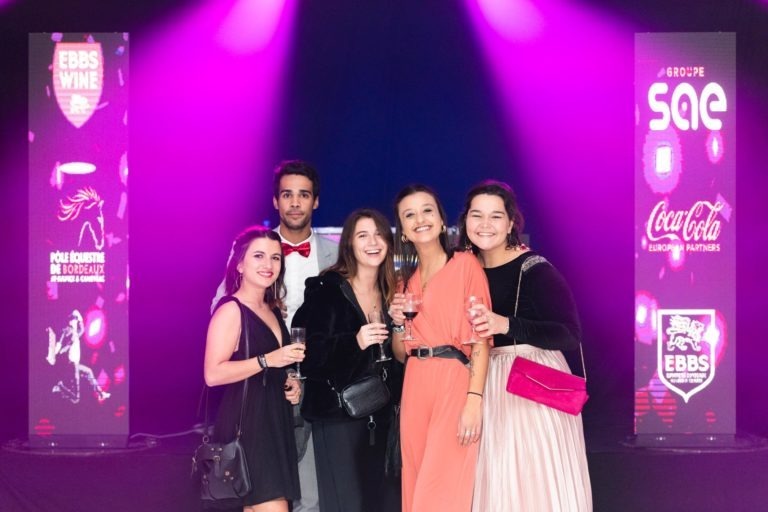 Un groupe de cinq personnes se tient debout ensemble lors d'un événement, souriant et tenant des verres à boire. Deux bannières verticales avec des logos, notamment EBBS Wine, Groupe SAE et Coca-Cola, sont visibles sur un fond sombre avec des lumières roses et violettes. Ils semblent profiter de la Cérémonie de remise des diplômes EBBS.