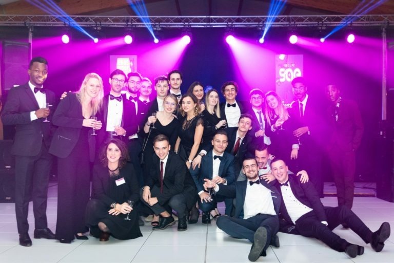 Un groupe d'une vingtaine de personnes bien habillées posent ensemble sur une piste de danse sous les lumières violettes de la scène lors de la cérémonie de remise des diplômes EBBS. La plupart sont en tenue de soirée, notamment en costume et en robe, certains tenant des verres. L'arrière-plan présente des équipements d'éclairage événementiel. Tout le monde sourit et semble passer un bon moment.