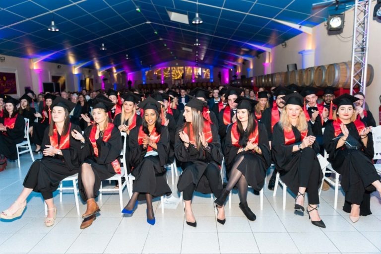 Un grand groupe d'étudiants est assis à l'intérieur lors de la cérémonie de remise des diplômes de l'EBBS, tous portant des casquettes et des robes noires avec des étoles rouges. Ils applaudissent et sourient, face à la scène. La salle a un plafond éclairé en bleu et un éclairage tamisé, avec des tonneaux décoratifs bordant les murs en arrière-plan.