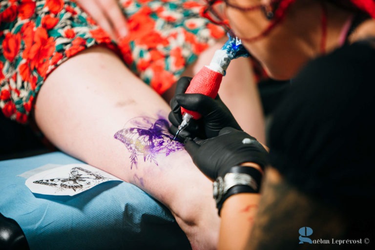 Au Salon Love Tattoo de La Teste-de-Buch, un tatoueur portant des gants noirs et un bandana rouge tatoue un papillon sur la jambe d'une personne. La personne, vêtue d'une robe à motifs floraux, a le contour visible sur sa peau avec quelques zones déjà ombrées. Un pochoir de papillon repose à proximité.