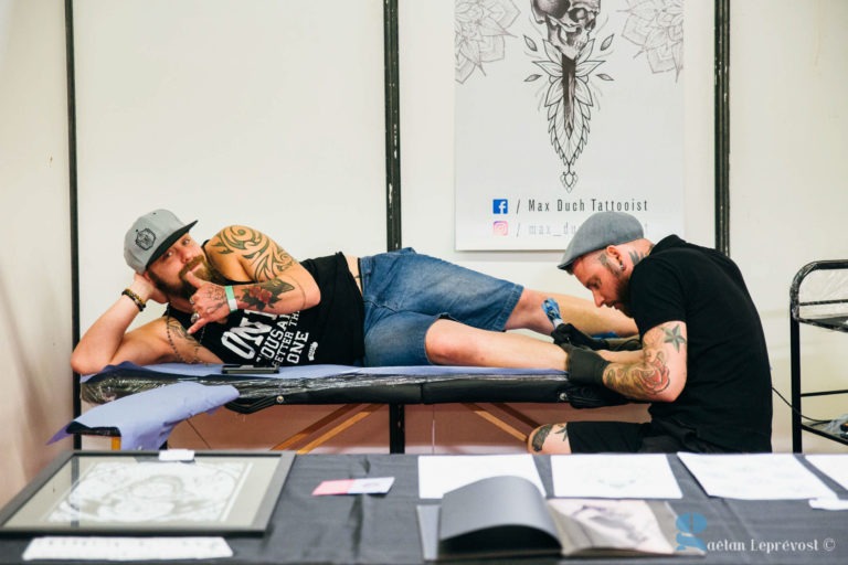 Au Salon Love Tattoo de La Teste-de-Buch, un tatoueur coiffé d'un chapeau gris et de gants tatoue la jambe d'un homme. L'homme, également coiffé d'un chapeau gris, est allongé sur la table, le sourire aux lèvres, le bras derrière la tête. L'arrière-plan présente divers motifs de tatouage et illustrations, dont un grand dessin présentant les coordonnées de l'artiste.