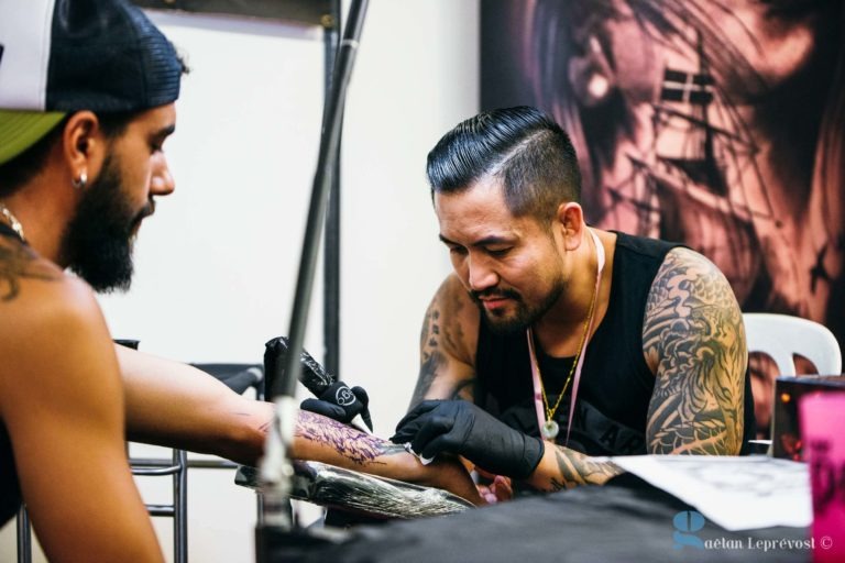 Un tatoueur portant des gants noirs, un débardeur noir et des tatouages sur les bras se concentre sur le tatouage de l'avant-bras d'un homme assis avec une barbe et une casquette au Salon Love Tattoo La Teste-de-Buch. Le bras du client repose sur une surface rembourrée, tandis que l'arrière-plan comprend un décor sur le thème du tatouage. Le crédit photo est visible en bas à droite.