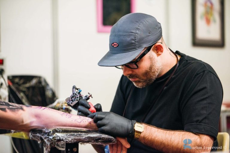 Un tatoueur du Salon Love Tattoo de La Teste-de-Buch, vêtu d'un t-shirt noir, de gants noirs et d'une casquette, se concentre pendant qu'il tatoue le bras d'une personne. Il tient une machine à tatouer dans sa main droite et travaille avec diligence. Derrière lui se trouvent divers outils et des œuvres d'art encadrées sur le mur. L'ambiance de la pièce est professionnelle et artistique.