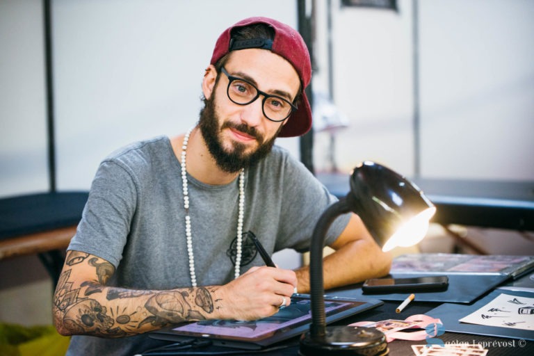 Un homme barbu avec des lunettes et coiffé d'une casquette rouge à l'envers est assis à une table du Salon Love Tattoo, à La Teste-de-Buch. Il dessine ou travaille sur un motif sous une lampe de bureau noire. Vêtu d'un t-shirt gris, il a des tatouages sur le bras droit. Divers papiers et outils sont étalés devant lui.