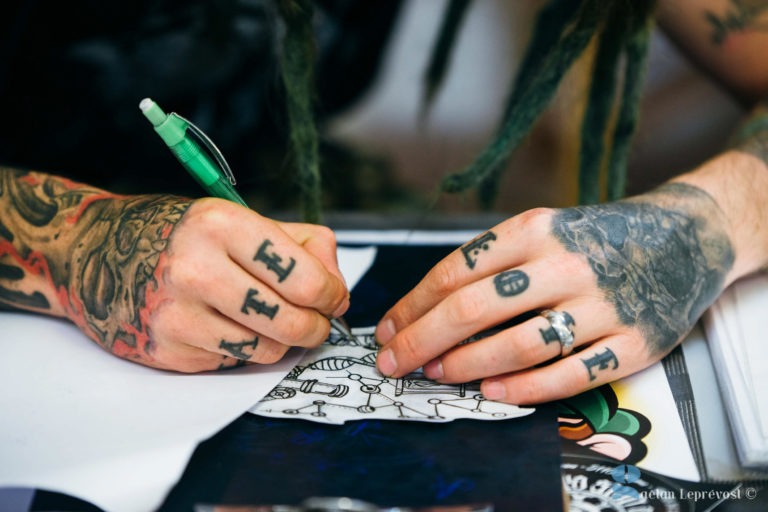 Une personne tatouée sur les deux mains, dont les lettres « LATE » et « LOVE », dessine sur un morceau de papier avec un stylo vert au Salon Love Tattoo de La Teste-de-Buch. Les bras de la personne sont également tatoués et elle se concentre sur le dessin. Des dreadlocks sont visibles, recouvrant partiellement la main qui dessine.