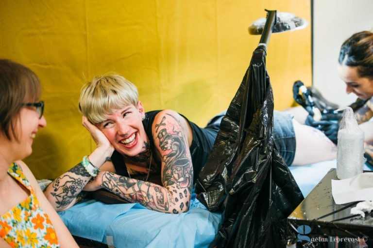 Une femme blonde tatouée sur les bras et les épaules rit, allongée sur une table du Salon Love Tatoo La Teste-de-Buch. Elle pose sa tête sur une main, vêtue d'un short. Un tatoueur travaille sur sa jambe tandis qu'une autre personne, souriante et portant des lunettes, est assise à proximité. Le fond est jaune.