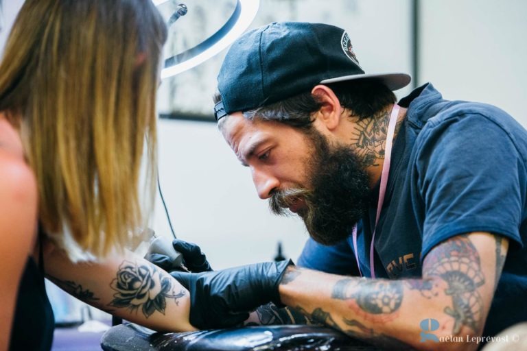 Un tatoueur barbu et tatoué se concentre sur le tatouage du bras d'une femme au Salon Love Tatoo de La Teste-de-Buch. La femme a un tatouage de rose et l'artiste porte des gants noirs, une casquette noire à l'envers et un t-shirt foncé. Une lumière est placée au-dessus d'eux, éclairant la zone de travail.