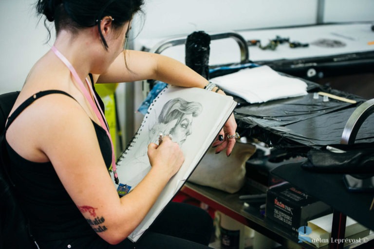 Une femme aux cheveux noirs, portant un haut noir et un cordon rose, est assise et dessine un portrait détaillé du visage d'une personne dans son carnet. Ses bras tatoués se déplacent gracieusement au milieu de l'espace de travail encombré rempli d'outils de dessin, d'une serviette blanche et de diverses fournitures artistiques rappelant le Salon Love La Teste-de-Buch.