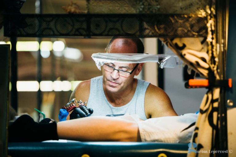 Au Salon Love Tattoo de La Teste-de-Buch, un tatoueur en chemise sans manches se concentre sur le tatouage du bas de la jambe d'un client allongé. L'artiste, tenant une machine à tatouer à deux mains, travaille au milieu de divers équipements et éclairages. Avec des lunettes sur les yeux, l'ambiance est professionnelle et concentrée.