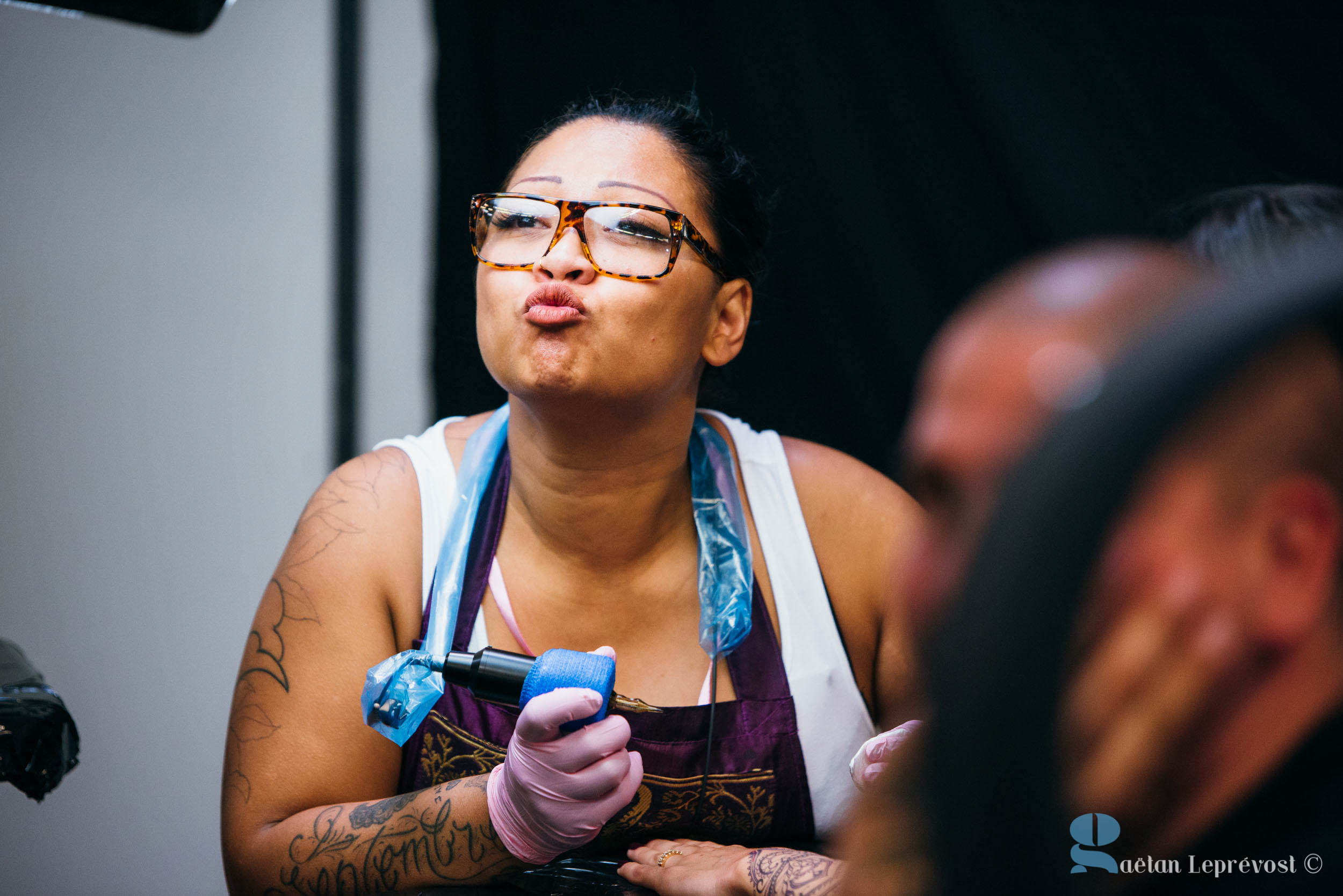 Une personne au bras tatoué, portant des lunettes et un haut sans manches, tient une machine à tatouer. Elle envoie un baiser ou fait une expression enjouée. L'arrière-plan est légèrement flou, d'autres personnes ne sont que partiellement visibles à l'intérieur du Salon Love Tattoo à La Teste-de-Buch. L'image porte un filigrane indiquant "Gaelan Leprevost ©".