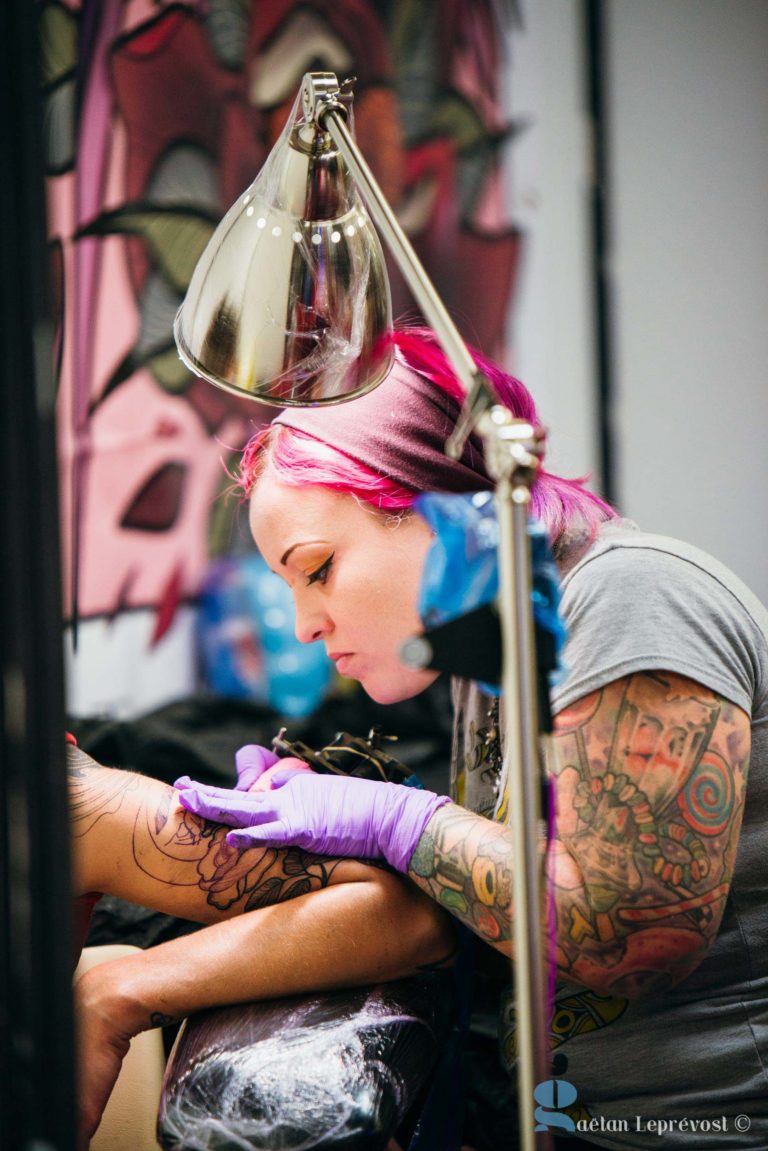 Un tatoueur aux cheveux roses, vêtu d'un t-shirt gris et de gants violets, travaille minutieusement sur un tatouage sur le bras d'une cliente au Salon Love Tattoo de La Teste-de-Buch. Le bras de l'artiste présente divers tatouages colorés. Une lampe éclaire la zone de travail et une fresque murale détaillée est visible en arrière-plan.