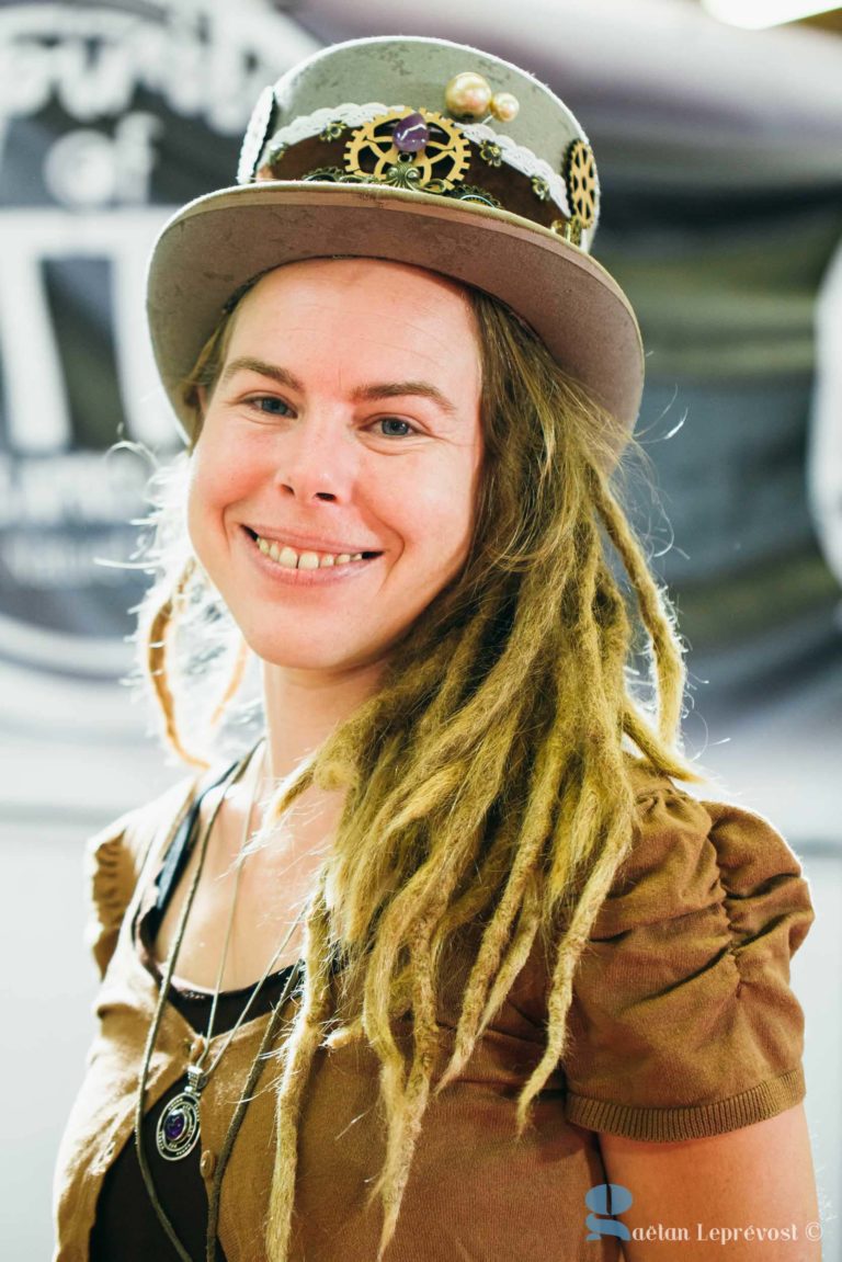 Une personne souriante aux longues dreadlocks porte un haut-de-forme marron décoré d'engrenages et d'embellissements. Elle est vêtue d'une tenue marron et d'un collier pendentif. L'arrière-plan est flou, avec une enseigne noire et blanche partiellement visible du Salon Love Tatoo La Teste-de-Buch. Le crédit photo est le suivant : "Gaetan Lepré vost.