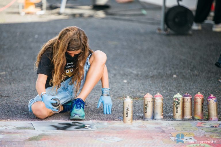 Une personne aux cheveux longs peint un pochoir sur le sol. Elle porte une chemise noire, des gants bleus et un masque. Diverses bombes de peinture sont placées à côté de sa zone de travail. La scène semble se dérouler en extérieur dans la ville ensoleillée de La Teste-de-Buch, dans une atmosphère généralement décontractée et créative.