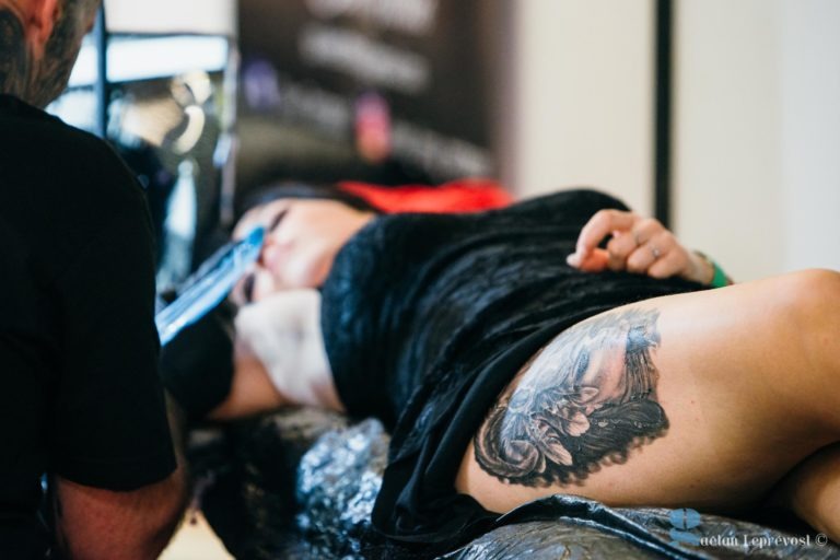 Une personne est allongée sur une table de tatouage au Salon Love Tattoo La Teste-de-Buch, recevant un dessin détaillé à l'encre noire sur le haut de sa jambe. Le tatoueur, dont le visage n'est pas visible, travaille méticuleusement. La personne porte une tenue noire, entourée de divers équipements de tatouage à l'intérieur.