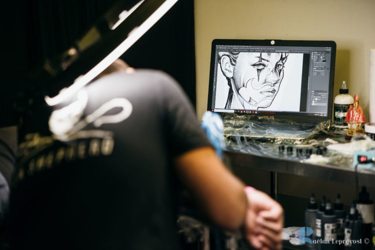 Un tatoueur se penche en avant, concentré sur son travail, dos à la caméra. Devant lui, un écran d'ordinateur affiche le croquis numérique du visage d'une femme avec des détails complexes à l'encre. Du matériel de tatouage et des encres sont disposés sur une table en métal à proximité. Le décor semble être le Salon Love Tattoo La Teste-de-Buch.