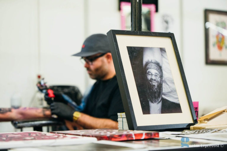 Un artiste portant une casquette et des lunettes se concentre intensément pendant qu'il tatoue une personne au Salon Love Tatoo La Teste-de-Buch. Au premier plan, on voit une photo en noir et blanc encadrée d'un homme avec une barbe et des lunettes. L'espace de travail est encombré de matériel de tatouage et de divers matériaux artistiques.