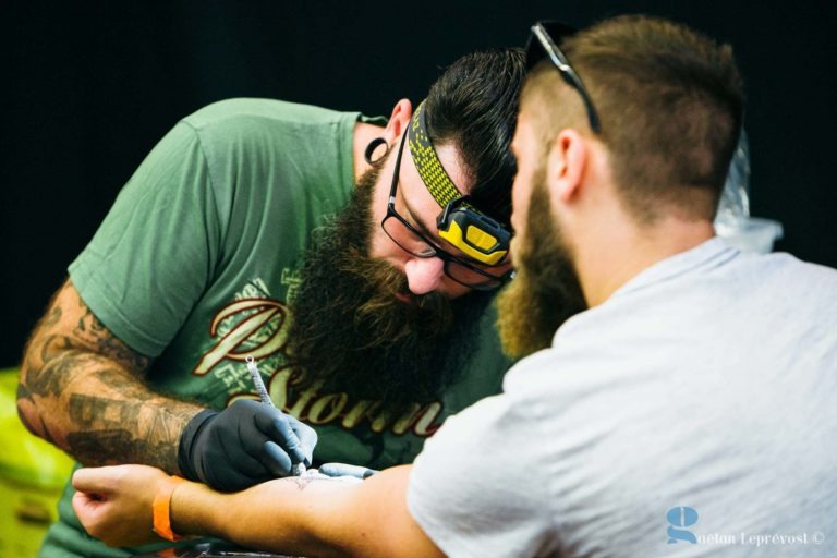 Un tatoueur barbu du Salon Love Tattoo de La Teste-de-Buch, muni d'une lampe frontale et de manches de tatouage, se concentre intensément sur l'encre du bras d'un client aux cheveux courts et portant une chemise grise. L'artiste, portant des gants noirs et un t-shirt vert à motifs, travaille méticuleusement pendant que le client est assis, immobile, observant son bras.