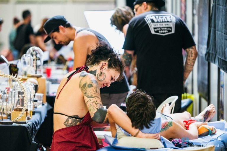 Au Salon Love Tatoo de La Teste-de-Buch, un tatoueur aux multiples tatouages et aux grandes boucles d'oreilles travaille sur le bras d'une personne dans le studio animé. Plusieurs autres artistes et clients sont visibles en arrière-plan, entourés de divers équipements de tatouage et de bouteilles d'encre. L'ambiance est concentrée et dynamique.