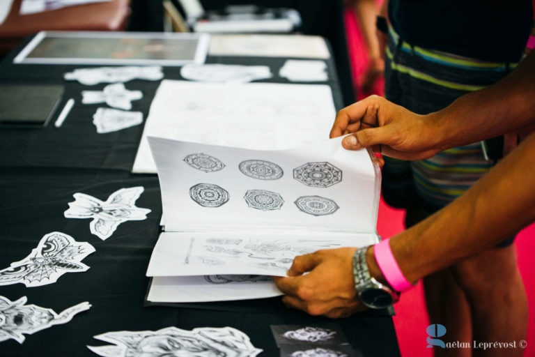 Une personne portant un bracelet rose feuillette un portfolio de dessins de tatouages posé sur une table recouverte d'une nappe noire au Salon Love Tatoo de La Teste-de-Buch. Divers dessins de tatouages, dont des mandalas et un papillon, sont étalés sur la table, évoquant l'effervescence d'une convention de tatouage ou d'un environnement de studio animé.