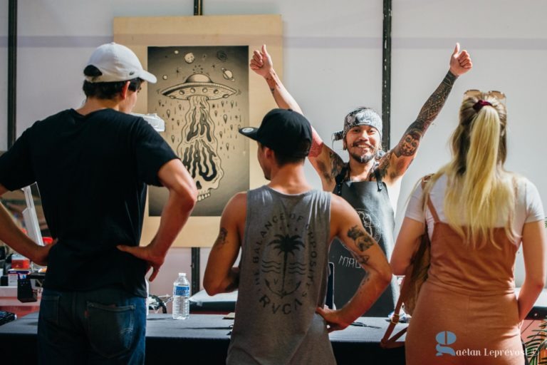 Derrière une table, un tatoueur aux bras tatoués et coiffé d'un bandana sourit et lève les pouces. Trois personnes se tiennent devant lui et discutent. Le fond du Salon Love Tatoo de La Teste-de-Buch est orné d'une œuvre d'art monochrome encadrée représentant une méduse et des ovnis, avec un filigrane indiquant « Gaetan Lepronovicz ».