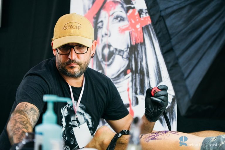 Un tatoueur du Salon Love Tattoo La Teste-de-Buch, coiffé d'une casquette de baseball beige et de gants noirs, se concentre sur le tatouage du bras d'un client. Derrière lui, on peut voir une œuvre d'art saisissante en noir et rouge représentant une femme portant un bâillon et un couvre-chef. Diverses fournitures de tatouage sont éparpillées un peu partout et un fond noir est visible derrière l'artiste.