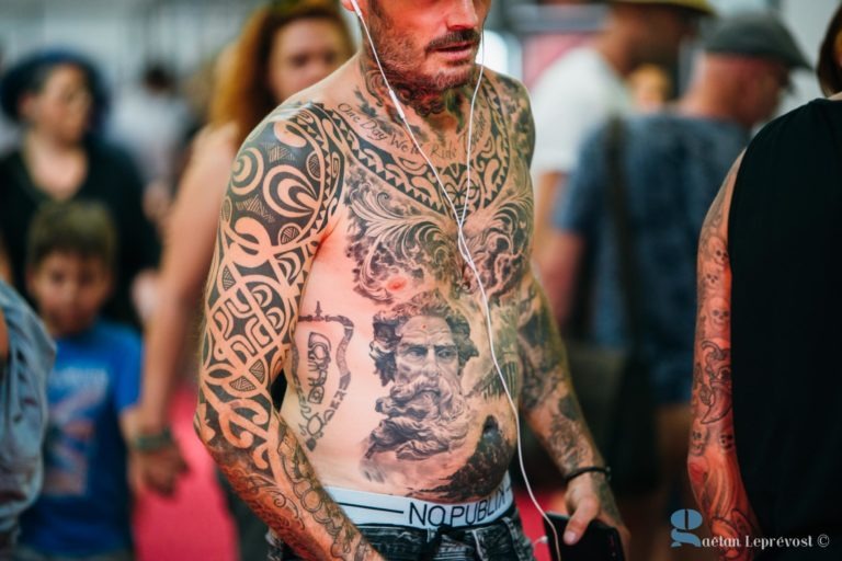 Un homme torse nu, avec des tatouages élaborés sur le corps, dont un grand tatouage de portrait sur le ventre, écoute de la musique avec des écouteurs. Il porte des sous-vêtements blancs, partiellement visibles au-dessus d'un pantalon foncé. Des gens sont à l'arrière-plan et la scène semble se dérouler dans un salon de coiffure à La Teste-de-Buch.