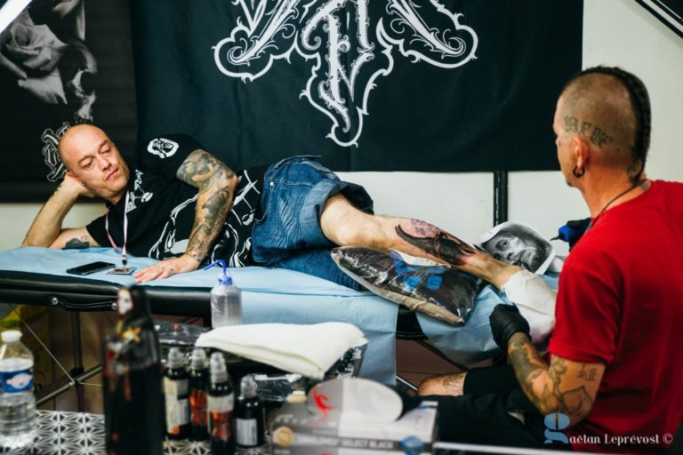 Un homme tatoué est allongé sur une table rembourrée au Salon Love Tatoo de La Teste-de-Buch, en train de se faire tatouer la jambe par un artiste. L'artiste, vêtu d'une chemise rouge et de gants, est concentré sur son travail. L'arrière-plan présente une bannière noire avec des motifs artistiques. Divers équipements et fournitures de tatouage sont visibles sur une table à proximité.