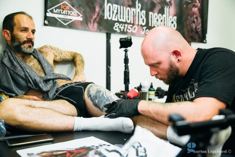 Un tatoueur, portant des gants et un t-shirt noir, travaille intensément sur un tatouage sur la jambe droite d'un client assis. Le client, portant une barbe et un foulard gris, est assis calmement. L'installation comprend des bouteilles d'encre et une machine. Une bannière en arrière-plan indique "Salon Love Tattoo La Teste-de-Buch 84750 Jo...".