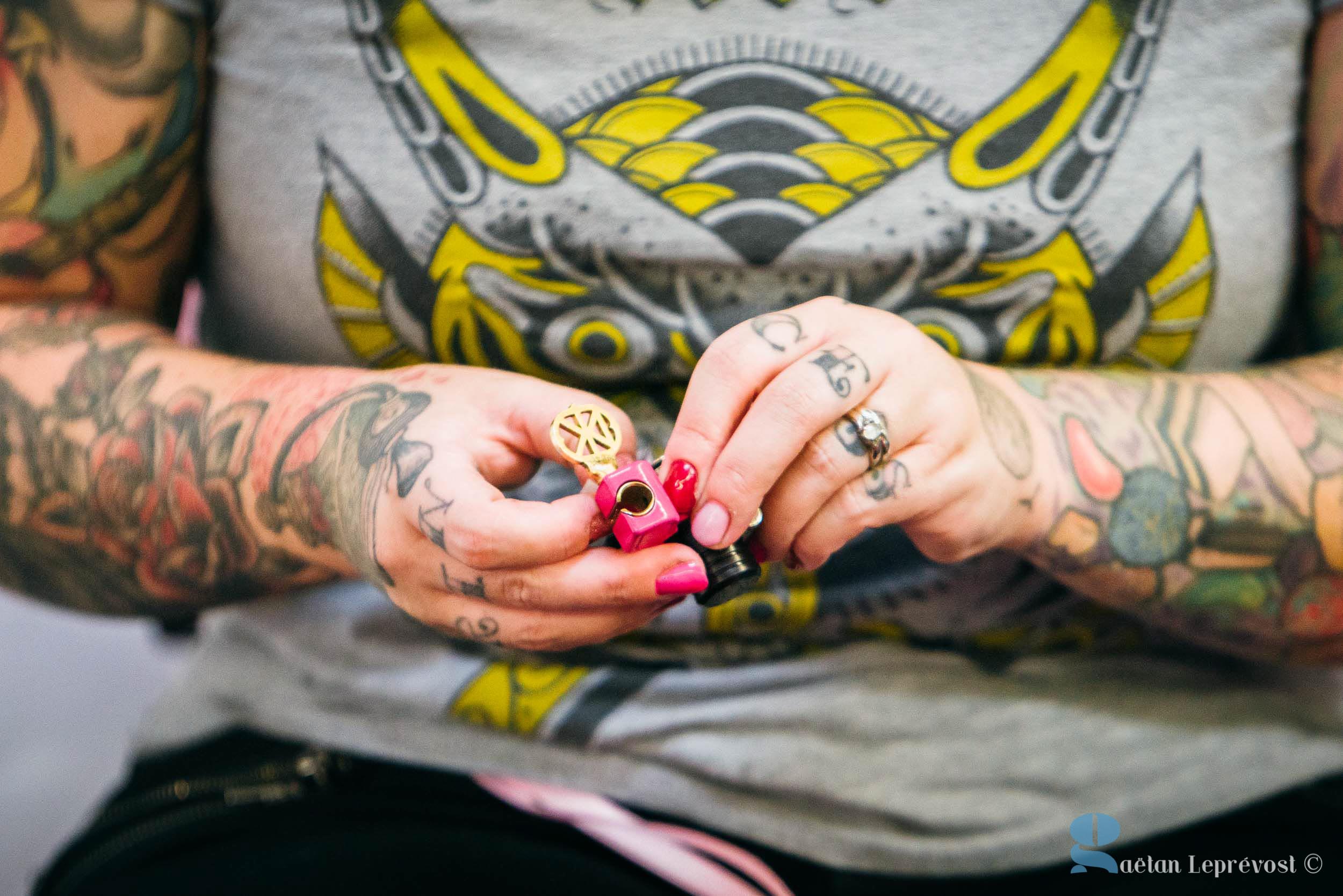 Une personne aux bras tatoués tient un petit objet complexe à deux mains. L'objet semble être jaune avec un motif, et un composant rose est en cours d'ajustement. Les ongles de la personne sont peints en rose et elle porte des bagues. Elle porte une chemise grise avec un graphique coloré sur lequel est écrit « Salon Love ».