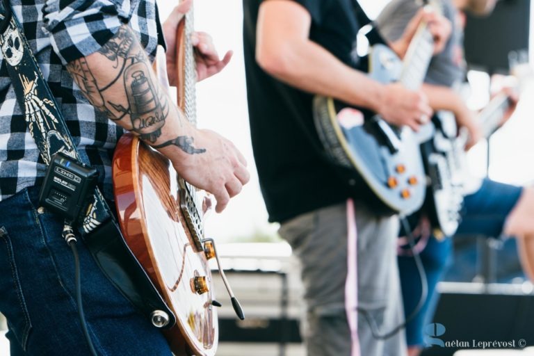 Gros plan sur deux musiciens jouant de la guitare électrique. Le guitariste de gauche, avec une chemise à carreaux et un avant-bras tatoué, porte un émetteur de guitare sans fil attaché à sa ceinture. Le guitariste de droite, en T-shirt noir uni, joue avec passion. Les deux guitares ont un design à corps solide, faisant écho au Salon Love Tattoo dans le style de La Teste-de-Buch.