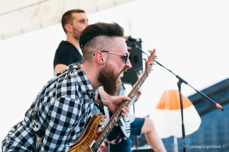 Un homme avec une barbe fournie et des lunettes de soleil joue passionnément de la guitare électrique sur une scène en plein air à La Teste-de-Buch, vêtu d'une chemise à carreaux noirs et blancs. À proximité, un autre musicien aux cheveux courts et à la chemise noire joue de la basse. Un pied de micro et un parapluie orange et blanc sont visibles.