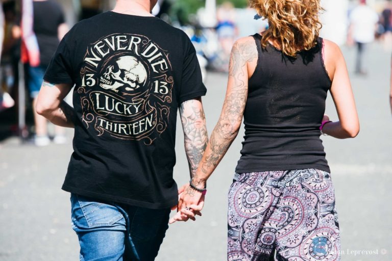 Un couple marche main dans la main dans une rue de La Teste-de-Buch. La personne à gauche porte un t-shirt noir avec un motif de tête de mort et le texte « Never Die Lucky 13 ». L'autre personne porte un débardeur noir et un pantalon à motifs. Tous deux ont des tatouages sur les bras, mettant en valeur l'art de Salon Love Tattoo, et affichent une tenue décontractée et estivale.