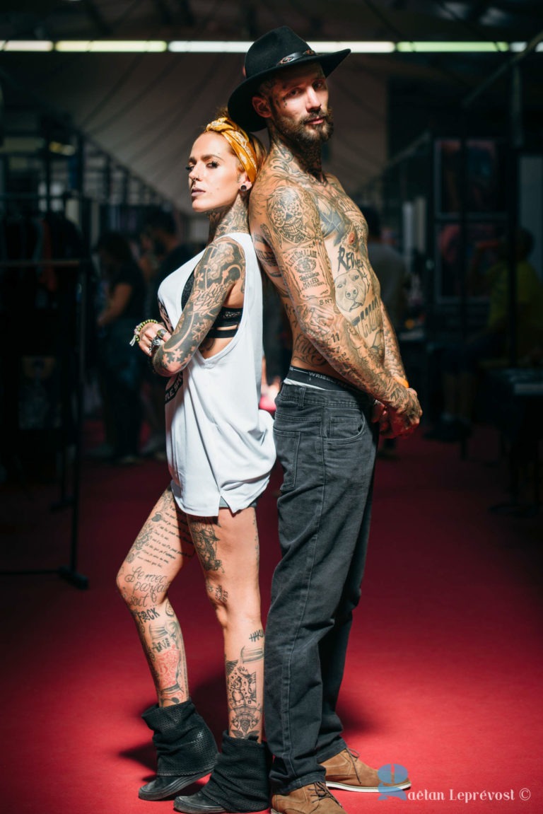 Deux individus lourdement tatoués se tiennent dos à dos dans un décor intérieur bien éclairé. La femme de gauche, aux cheveux blonds courts noués dans un bandana, porte un haut blanc sans manches, un short noir et des bottes. L'homme de droite, portant une barbe et un chapeau noir, est torse nu avec un pantalon gris et des bottes noires. Leurs tatouages complexes font allusion à leur amour commun pour l'art à l'encre de Chine.