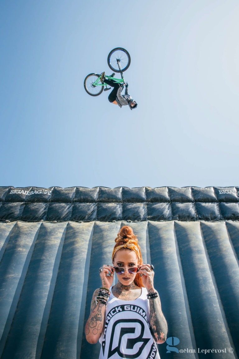 Une personne portant un foulard orange et des lunettes de soleil, ornée de plusieurs bracelets et tatouages du Salon Love Tatoo de La Teste-de-Buch, pose avec assurance au premier plan. Ci-dessus, un cycliste exécute une impressionnante cascade en plein air devant un grand coussin gonflable sous un ciel bleu dégagé. Photo de Gaëtan Leprévost.