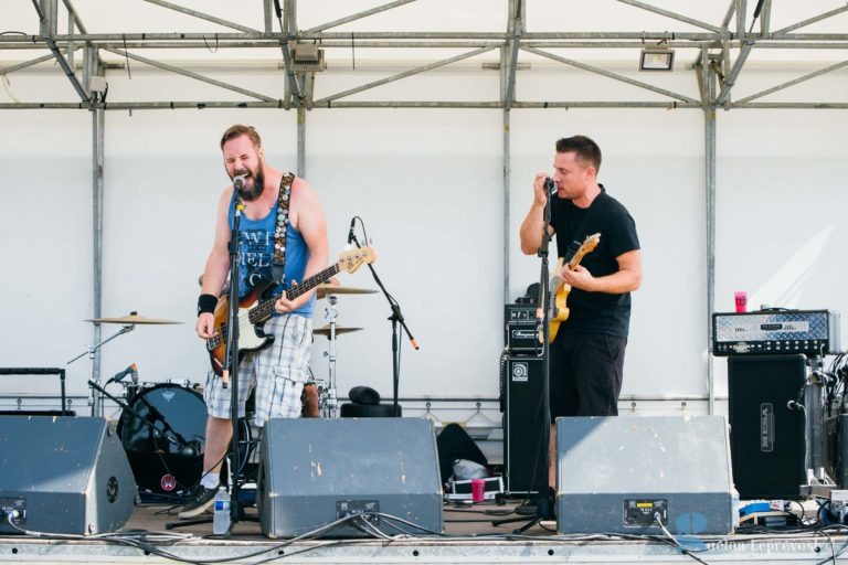 Deux musiciens se produisent sur une scène en plein air à La Teste-de-Buch. Le chanteur de gauche, vêtu d'un débardeur bleu et d'un short à carreaux, joue de la guitare basse avec énergie. Le guitariste de droite, vêtu de noir, chante dans un micro. Des tambours et d'autres équipements sont visibles en arrière-plan.